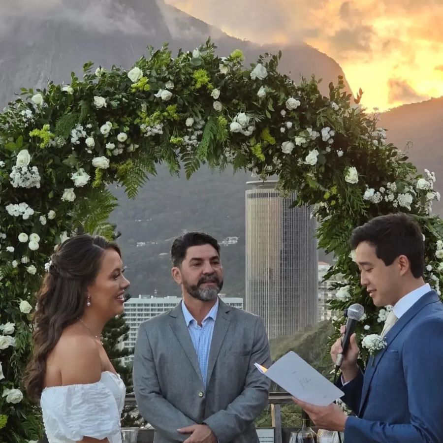 Laura e Fábio sob o arco de flores ao pôr do sol, com a cidade ao fundo
